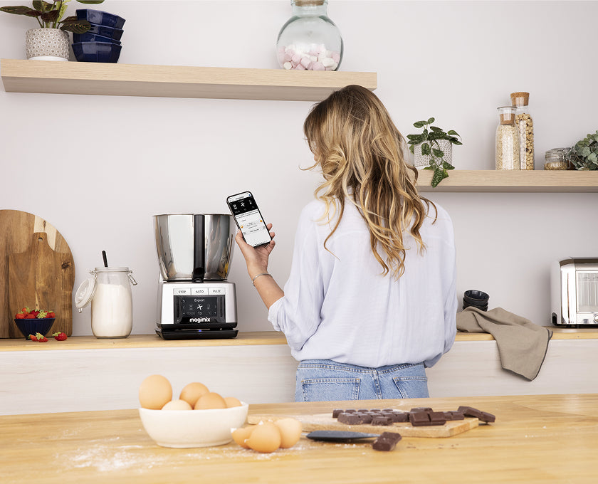 Une femme dans sa cuisine pilote son robot cuiseur connecté depuis son smartphone
